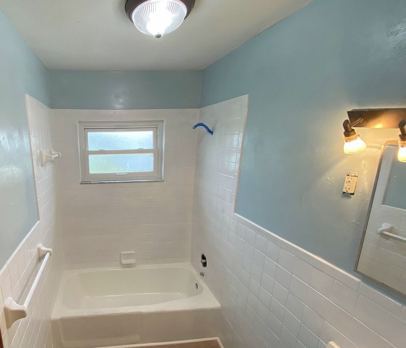 The same bathroom afterward: a white tub and tile, with freshly painted pale blue walls.