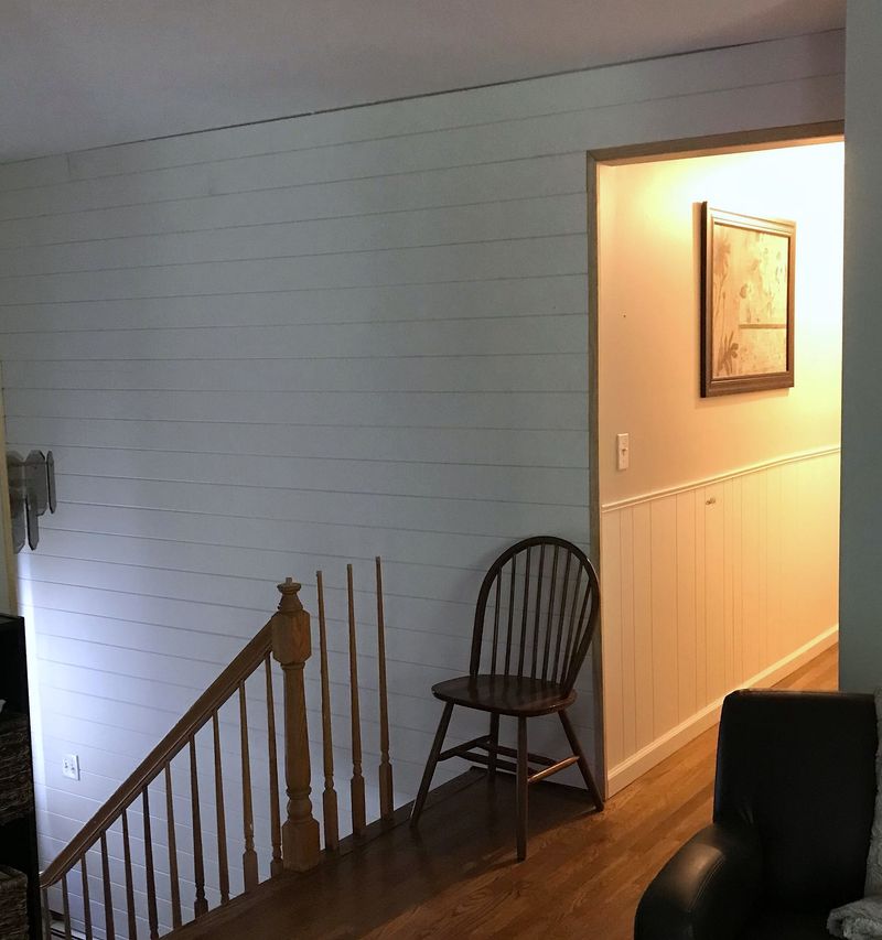 A stair landing with a white painted shiplap wall, a wood railing, and a chair beside a lit doorway.