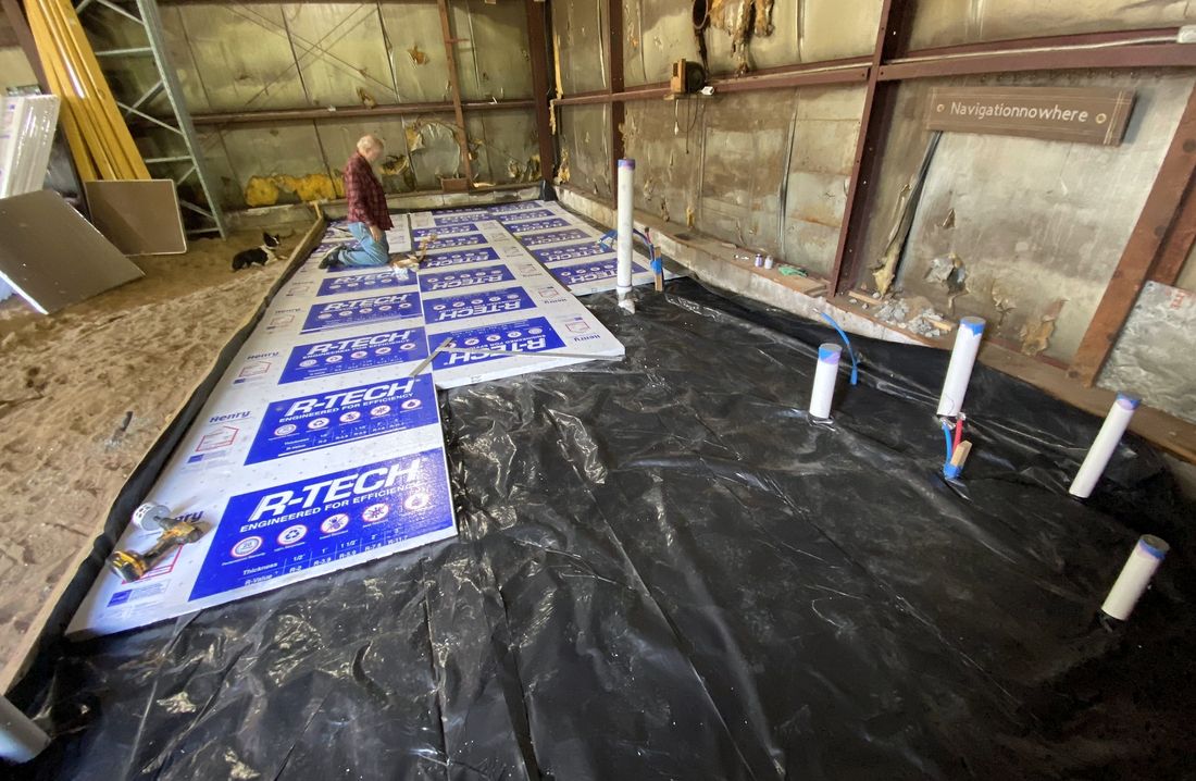 Insulation board and a black vapor barrier laid over gravel inside a building, ahead of a concrete slab.